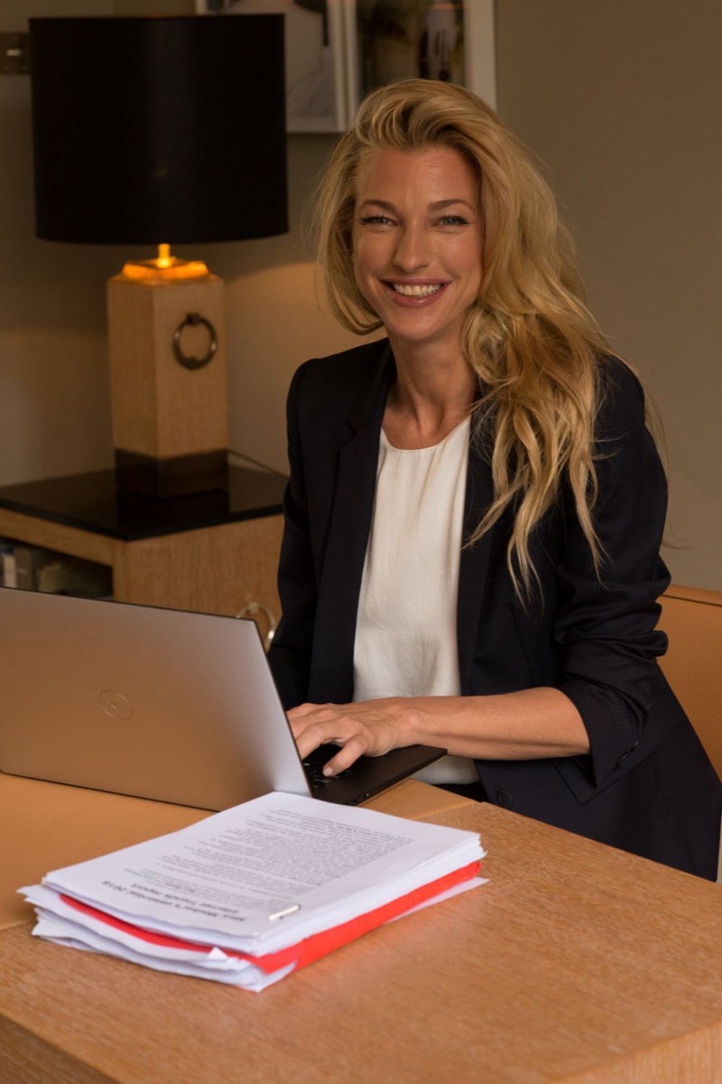Retrato de Nicole Junkermann con blazer azul marino, sonriente, trabajando con su portátil en un escritorio iluminado